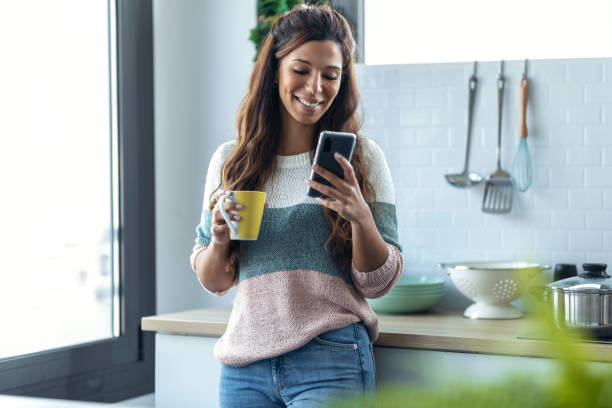 istock Beautiful young woman using her mobile phone while drinking a cup of coffee in the kitchen at home. 1309887424