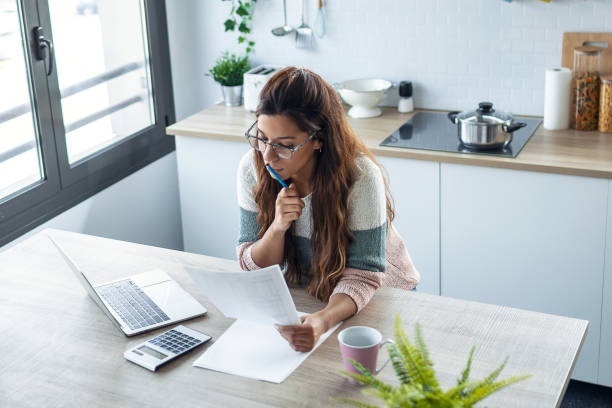 istock Pretty young business woman working with computer while consulting some invoices and documents in the kitchen at home. 1309887780