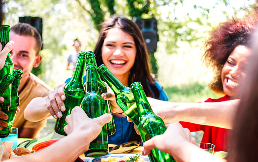Happy millenial friend having fun at barbecue garden party - Life style and friendship concept with young people toasting bottled beer at summer hangout - Warm bright filter with focus on bottles