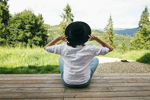Young female in hat and casual cloth sitting on porch on background of sunny mountains hills. Stylish hipster woman relaxing on wooden terrace. Calm tranquil moment. Travel and wanderlust