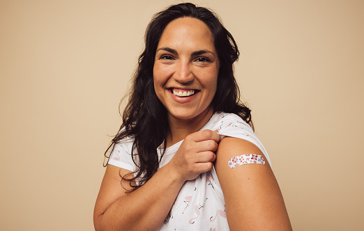 Portrait of a female smiling after getting a vaccine. Woman holding up her sleeve and showing her arm with bandage after receiving vaccination.