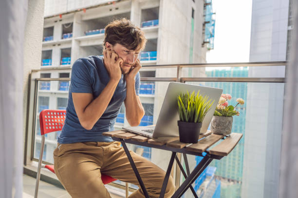 le jeune homme essayant de travailler sur le balcon ennuyé par le bâtiment travaille dehors. concept de bruit. pollution atmosphérique causée par la poussière des bâtiments - toughness endurance built structure architectural styles photos et images de collection