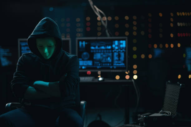 istock Wanted Hacker with Masked Face looks into the Camera. In the Background His Operating Room with Multiple Displays and Cables. After a while he turns away from the camera and goes back to work 1309899351