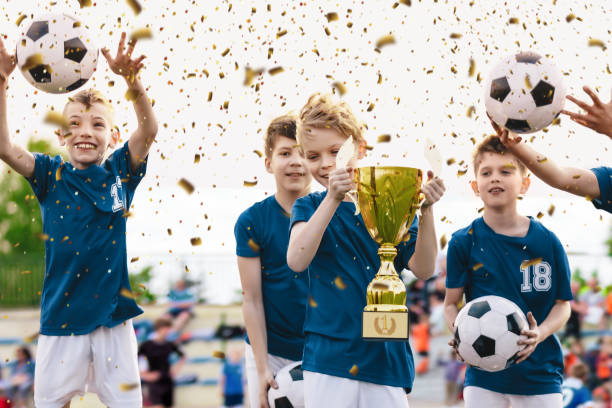 soccer team celebration. cheerful children celebrating success in football tournament game. junior sports team rising golden trophy on confetti celebration. boys jumping and throwing balls in joy - soccer celebration success group of people imagens e fotografias de stock