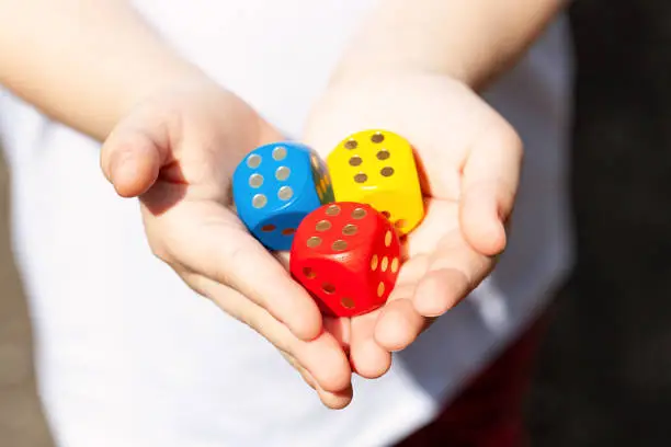 Photo of Little child holding three colorful wooden dice showing six in her small hands, closeup. Multi colored game dice with number 6 on them. Fortune, success, winning throw luck abstract concept, good odds