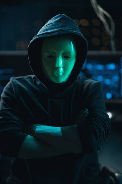 istock Dangerous Internationally Wanted Hacker with Masked Face Speaks into the Camera. In the Background His Operating Room with Multiple Displays and Cables 1309909581