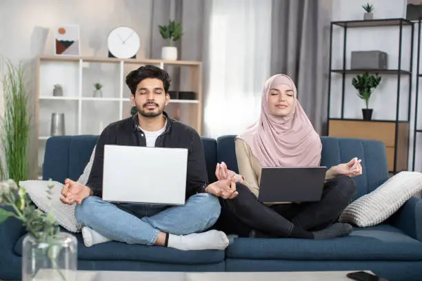 Photo of Pleasant likable young arabian muslim man and woman in hijab, sitting in lotus pose with laptop pc on sofa at living room, and meditating with closed eyes, folding fingers in mudra