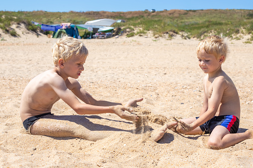 Little blond boys sit on the sand by the sea and play with the sand. Tent town in the background. Summer happy holidays.