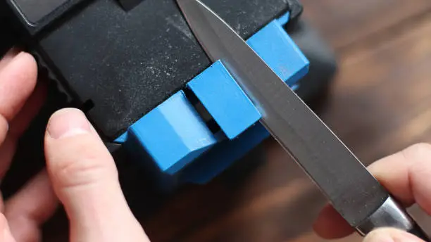 Sharpening a knife on an electric sharpener at home. The man's hand drives the knife blade between the blue sharpeners, dust flies on the machine.