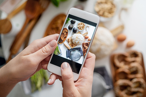 Food blogger taking a picture with her phone of groceries on white table. Picture of ingredients for breakfast on smartphone. Blogging concept.