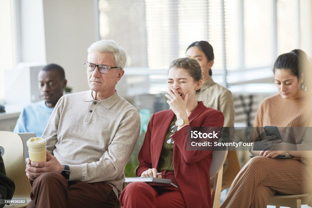 Young Woman Yawning at Business Seminar Diverse group of business people sitting on chairs in audience and listening at meeting or seminar, focus on young businesswoman yawning in foreground Boredom Stock Photo
