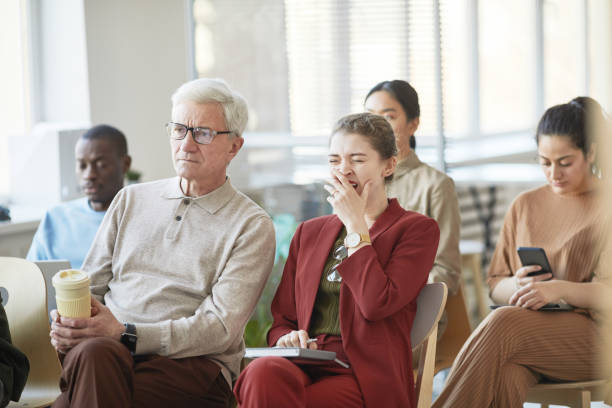 Young Woman Yawning at Business Seminar Diverse group of business people sitting on chairs in audience and listening at meeting or seminar, focus on young businesswoman yawning in foreground boredom stock pictures, royalty-free photos & images