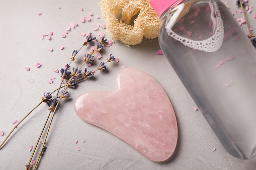 Rose quartz gua sha tool, dried lavender flowers and cosmetic product on grey table, closeup