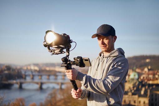 Young man filming with camera and gimbal. Filmmaker standing against urban skyline. Prague, Czech Republic