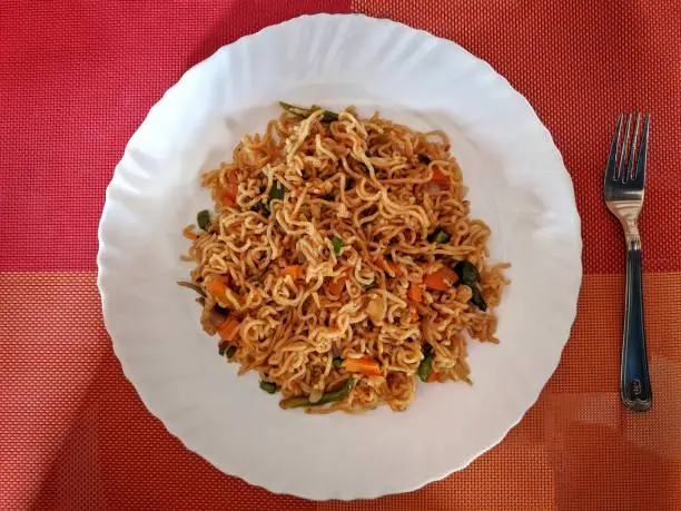 Hyderabad India - March 10, 2021: Vegetable Maggi Recipe | Tasty Veg Maggi | Veg Maggi Masala Noodles | Easy Quick Veg Noodles in a white plate beside the forks and spoon with colourfull background, india