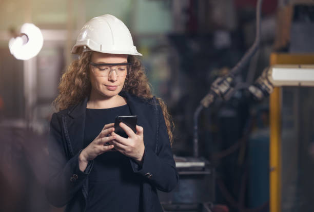istock Female Industrial Worker in the Hard Hat Uses Mobile Phone While Walking Through Heavy Industry Manufacturing Factory. 1309937154