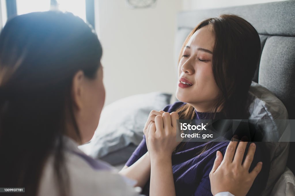 Female doctor giving consoling to a stressed exhausted upset female patient suffering from women’s sickness and emotional stress in bedroom Doctor explaining medical consultation to a stressed exhausted upset female patient during home visit. House call & Home Medical, Healthcare & Medicine Concepts. Healthcare Worker Stock Photo
