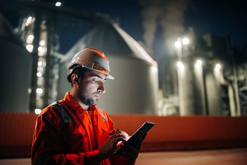 Caucasian man engineer using digital tablet working late night shift at petroleum oil refinery in industrial estate