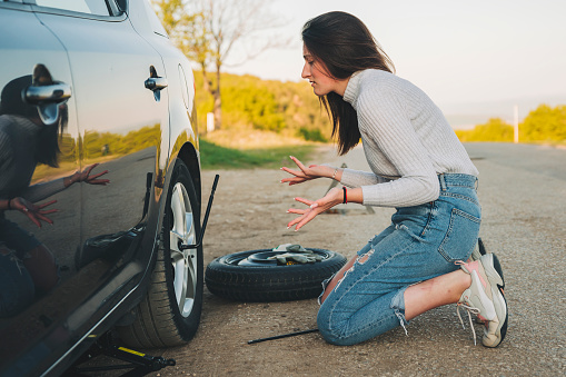 Woman changing wheel after a car breakdown at the side of the road. Transportation, traveling concept. She looks tired and desperate, and waiting help from someone