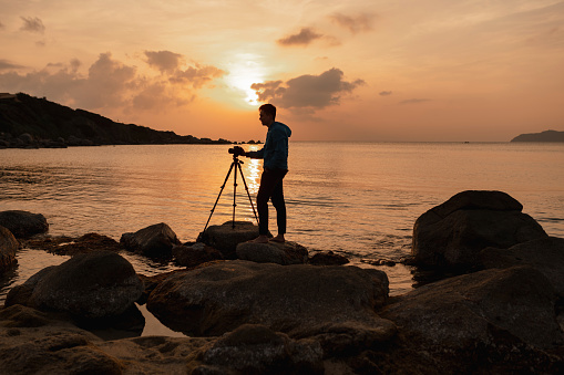 Silhouette of photographer preparing equipment to take a photo at the sunset with camera and tripod.