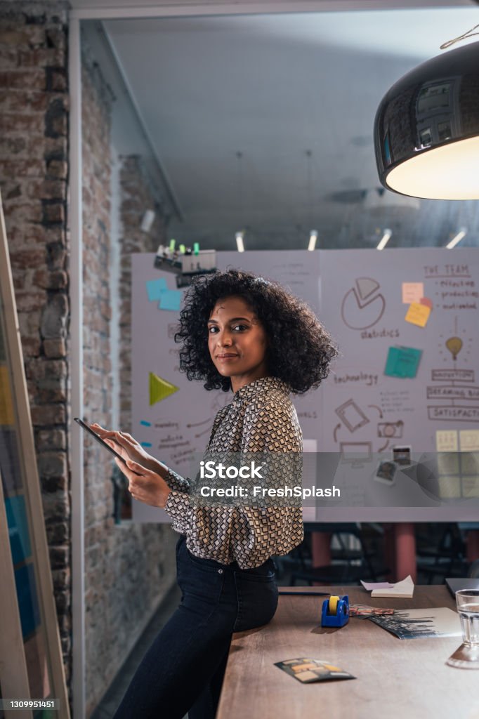 New Normal Concept: Side View of a Young African American Young Female Entrepreneur Holding Files, Alone at the Office Creative professional/ designer at work place, holding a tablet and files/documents, leaned against the desk. Business Stock Photo