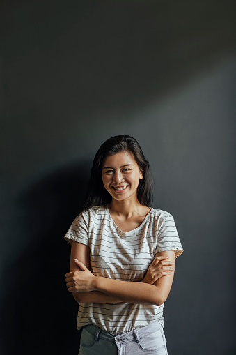 A portrait of a happy young woman in a white T-shirt with black stripes, standing against a dark gray background.