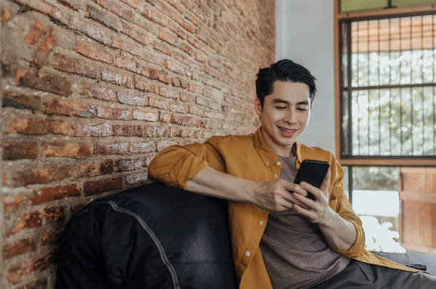 Happy Asian Student Sitting on the Sofa and Texting on his Mobile Phone A young smiling Asian man chatting on his smartphone and sitting in the living room. scrolling stock pictures, royalty-free photos & images