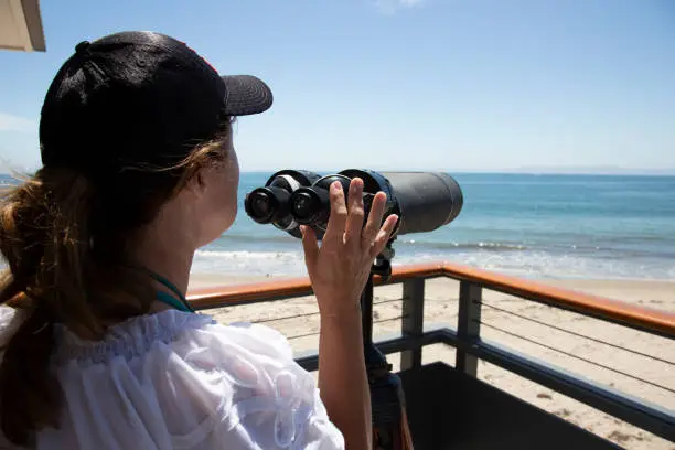 Woman with black baseball cap, brown hair in ponytail, and white blouse, has hand on binoculars while gazing out at the blue pacific ocean