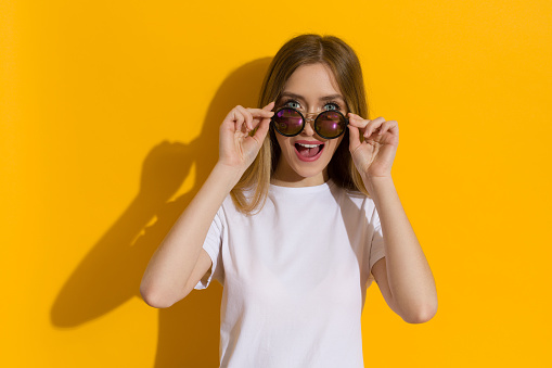 Happy young blond woman in white shirt is looking at camera over sunglasses and talking. Waist up shot on yellow background.