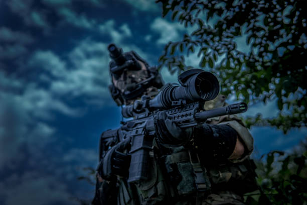 The soldier is holding a weapon with a telescopic sight. View from the lower angle. Night vision device in the dark.