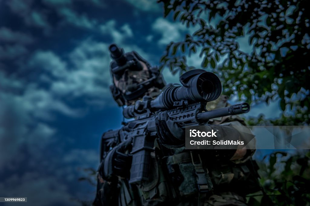 The soldier is holding a weapon with a telescopic sight. View from the lower angle. Night vision device in the dark. Night Vision Stock Photo