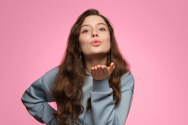 A beautiful girl with long hair sends an air kiss. A beautiful girl with long hair in a blue hoodie sends an air kiss on a pink background. blowing a kiss stock pictures, royalty-free photos & images