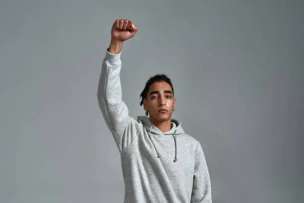 Portrait of young Romani man in hooded sweatshirt protesting against gray background.