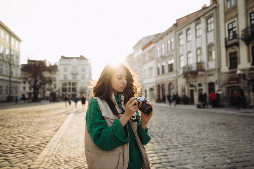 A cheerful woman taking a photo on an analogue camera in the old European city at the sunset