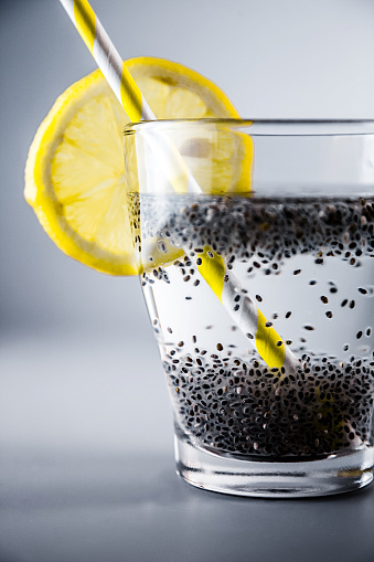 Studio shot of Chia seeds Lemon Water in glass cup with slice of lemon, drinking straw and spoon. Clean studio shot, neutral gray background. Yellow and gray contrast. Detox, healthy diet drink.