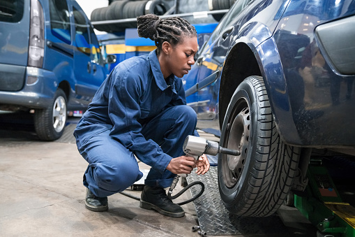 Auto mechanic or apprentice squatting next to the car in the auto repair shop. Young woman changing car wheel.