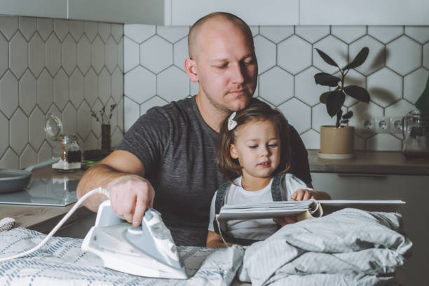 Man ironing bed linen and reading book to his little daughter. Father is engaged in household chores. Man ironing bed linen and reading book to his little daughter. Father engaged in household chores. laundry husband housework men stock pictures, royalty-free photos & images