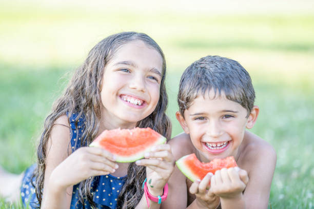 Two adorable siblings, a brother and sister are enjoying fresh watermelon outdoors on a summer day. They are at a water park and dressed in their bathing suit.