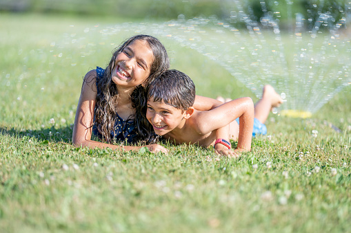 An adorable pair of brother and sister are lying down on green grass in their backyard. They have the water sprinklers on to beat the heat on a hot summer day. They are dressed in bathing suits. They are happy and smiling.