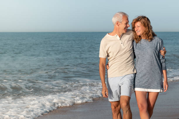 couples mûrs heureux marchant sur la plage dans les bras de l’un l’autre - senior adult women adult tan photos et images de collection