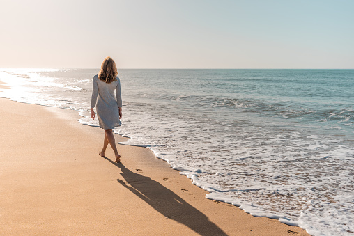 Woman walking towards the sunset light on the beach wearing a grey dress and barefoot