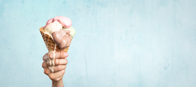 A hand holding an ice cream cone. The ice cream is dripping down as it melts. Isolated on a light blue background with copy space.