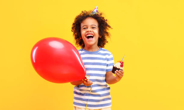 счастливый черный ребенок с воздушным шаром и день рождения кекс улыбается в желтой студии - party hat birthday celebration party стоковые фото и изображения