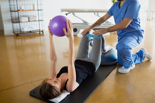 Female patient lying down on a mat, exercising with her physiotherapist using resistance band for legs and a fit ball, in a gymnasium or clinic. Physical therapy concept.