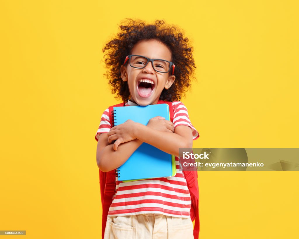 Cheerful little ethnic schoolchild with backpack and textbooks in yellow studio Positive little African American boy with curly hair in casual clothes and eyeglasses laughing while standing against yellow background with school backpack and copybooks Back to School Stock Photo