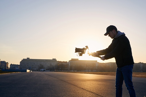 Young man filming with camera and gimbal. Videographer standing against city at sunrise. Prague, Czech Republic