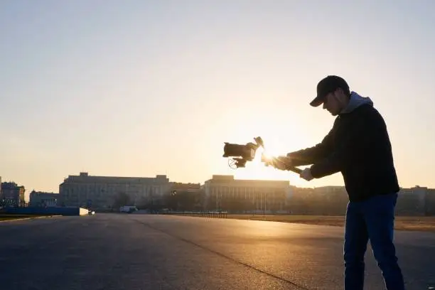 Young man filming with camera and gimbal. Videographer standing against city at sunrise. Prague, Czech Republic