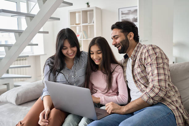 felice famiglia indiana con figlia bambina che si diverte usando il computer portatile a casa. genitori sorridenti e figlia adolescente che ridono guardando il dispositivo che naviga o guardano video divertenti seduti sul divano. - computer child family laptop foto e immagini stock