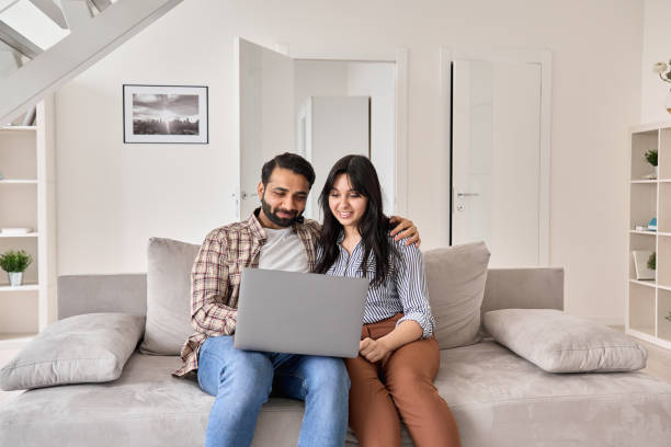istock Happy indian family couple using laptop looking at computer sitting on sofa together relaxing at home. Smiling ethnic husband and wife doing online ecommerce shopping on website in modern living room. 1310078151