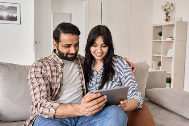 istock Happy indian family couple using digital tablet computer at home. Smiling young husband and wife watching tv, video calling or doing online ecommerce shopping together sitting on couch in living room. 1310078160